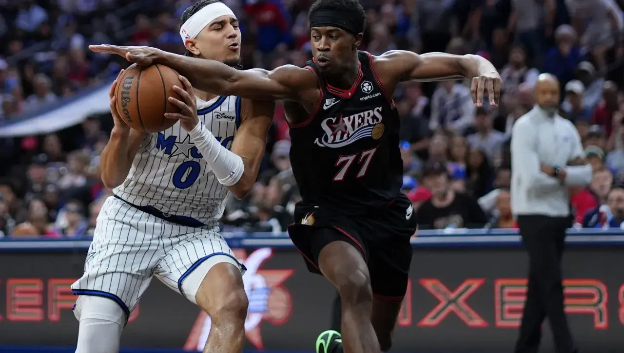 Two basketball players reach for the ball during a game: one in a white pinstripe uniform, the other in a black uniform with number 77, with a crowded arena in the background.