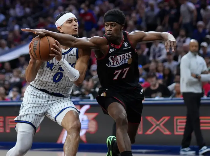 Two basketball players reach for the ball during a game: one in a white pinstripe uniform, the other in a black uniform with number 77, with a crowded arena in the background.