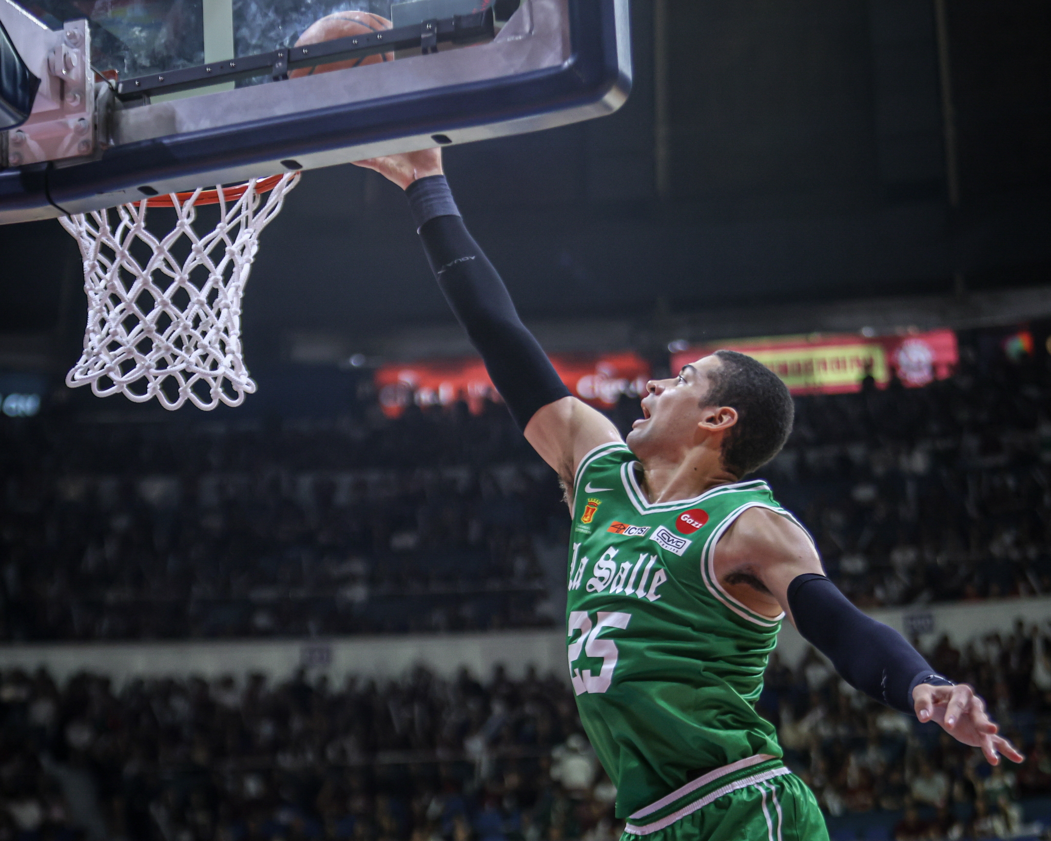 A photo taken from a low angle of Mike Phillips, wearing a green La Salle jersey with the number 25, in the middle of a dunk over the net. He has a determined and open expression and is looking upwards at the net. He is wearing long black compression sleeves on his arms. The background is a packed indoor arena.