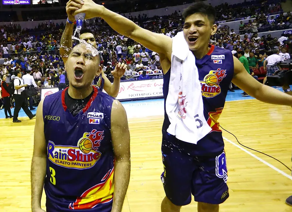 Two Rain or Shine players celebrate on a basketball court; one teammates sprinkles water over the other’s head while fans cheer in the background.