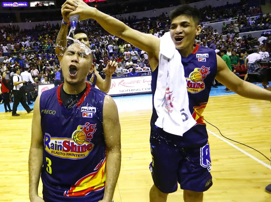 Two Rain or Shine players celebrate on a basketball court; one teammates sprinkles water over the other’s head while fans cheer in the background.