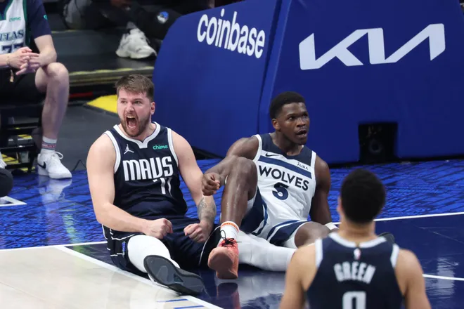 Two basketball players sit on the court after a play; one in a Mavs jersey shouts, the other in a Wolves jersey looks on.