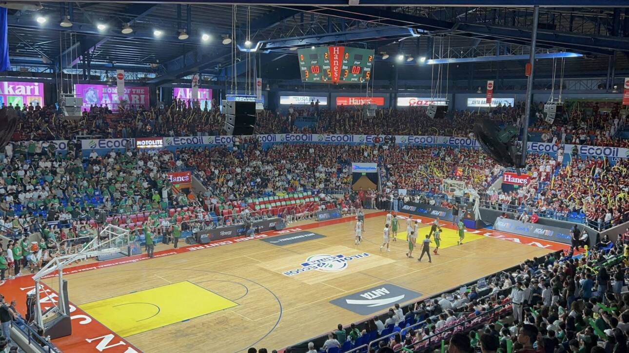 Indoor basketball arena filled with cheering fans; players on the court under bright lights.