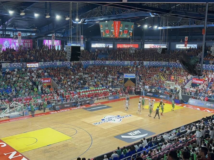 Indoor basketball arena filled with cheering fans; players on the court under bright lights.