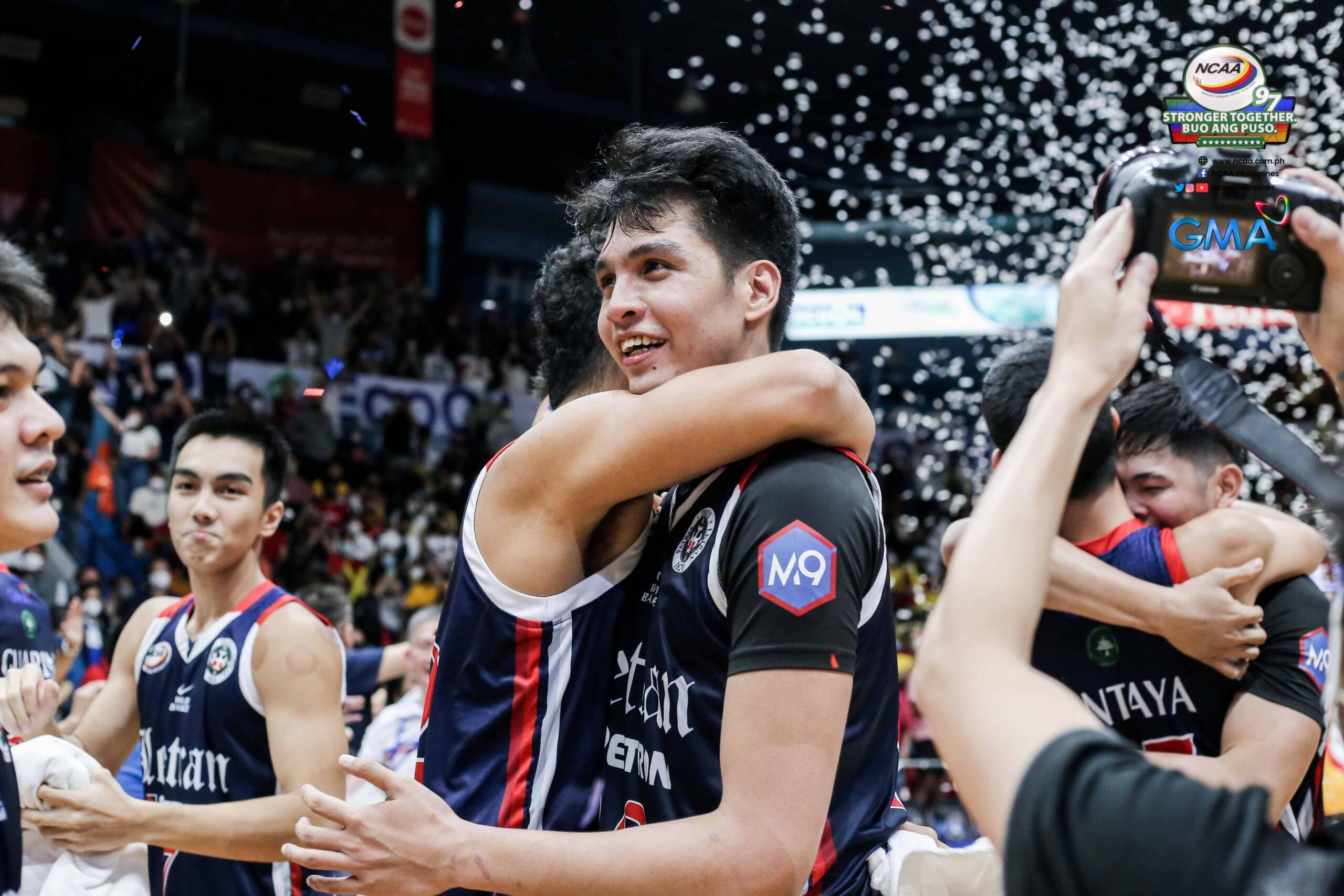 Basketball players in dark uniforms hug in celebration on the court as confetti falls and a cheering crowd looks on in the background.