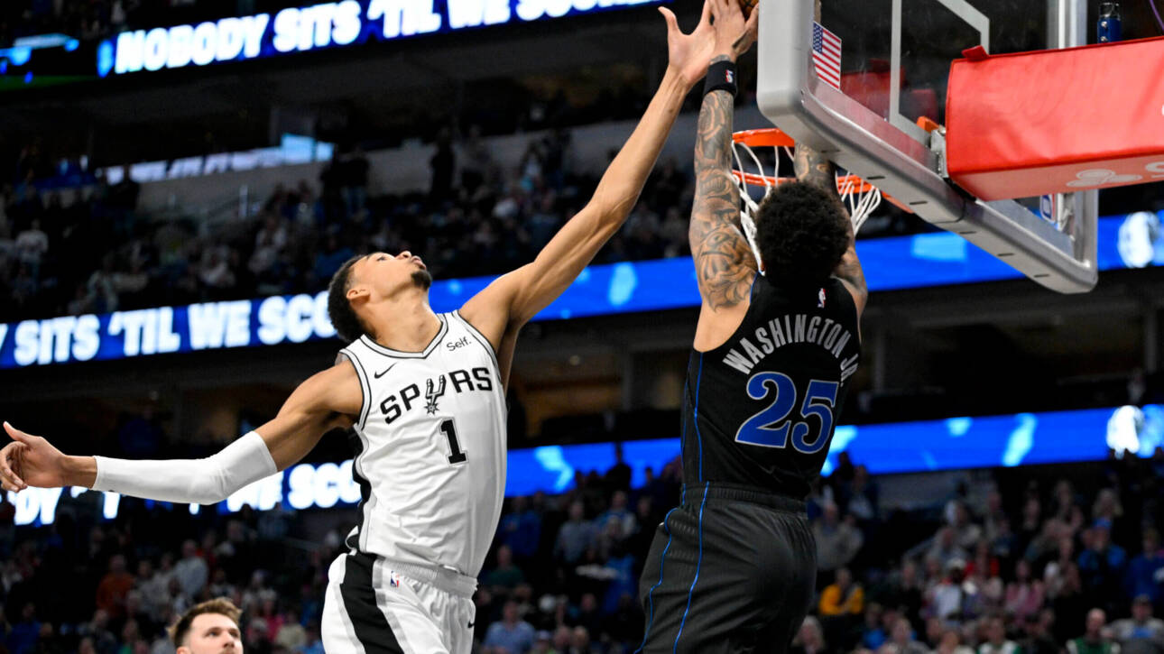 Two basketball players leap for the ball at the hoop, Spurs in white challenging Washington in dark uniform amid a crowded arena.