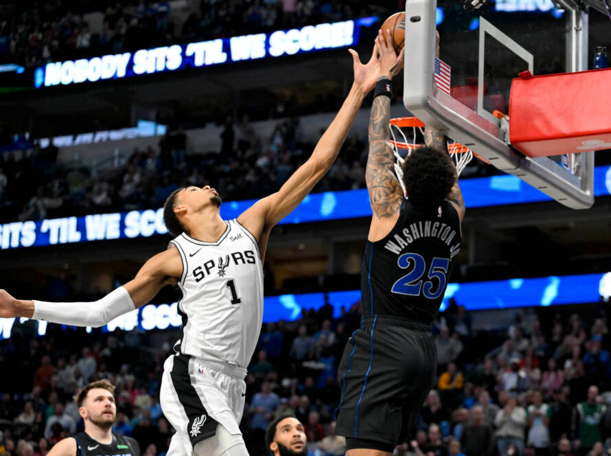 Two basketball players leap for the ball at the hoop, Spurs in white challenging Washington in dark uniform amid a crowded arena.