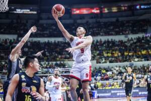 Scottie Thompson of Barangay Ginebra crashing the offensive glass against TNT defenders | GGSLOT | Sports & Games News Basketball player in white jersey (#9) leaps for a layup near the hoop as defenders reach; crowded arena in the background.
