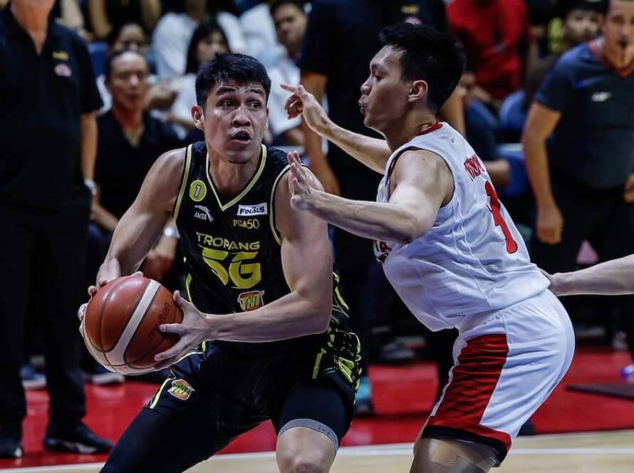 Basketball player in a black-yellow uniform drives with the ball as a defender in white-red blocks him in a crowded arena.