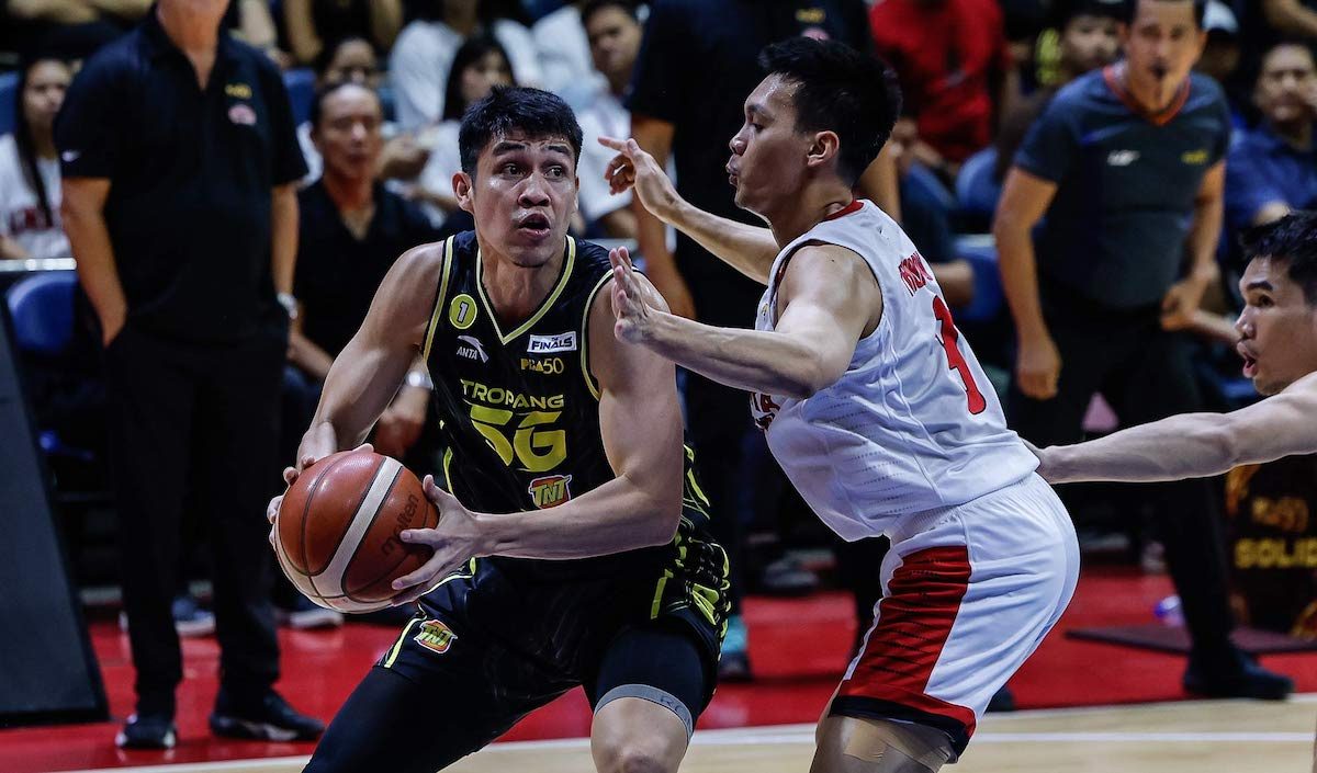 Basketball player in a black-yellow uniform drives with the ball as a defender in white-red blocks him in a crowded arena.