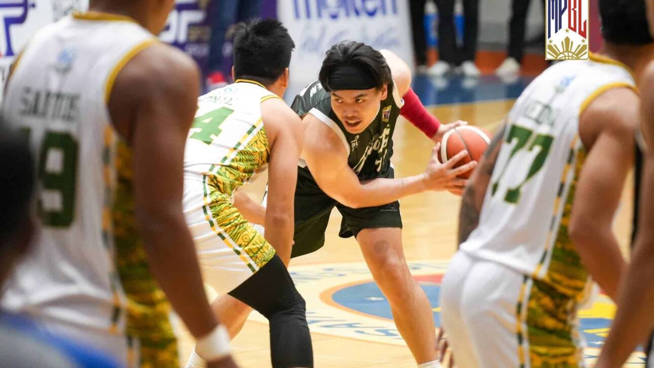 Player in a black jersey dribbles the ball as defenders in white-and-green converge on a crowded indoor basketball court.