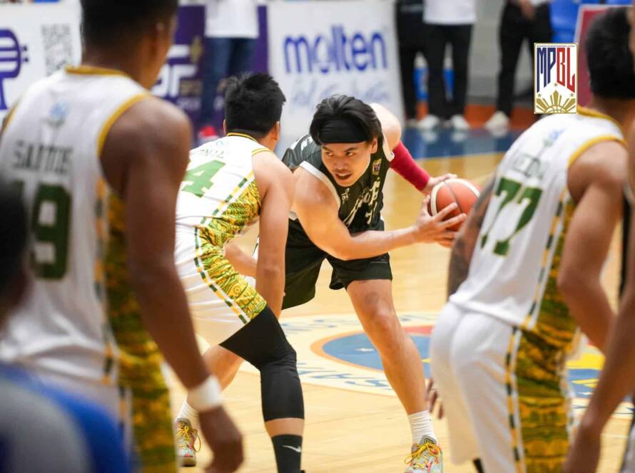 Player in a black jersey dribbles the ball as defenders in white-and-green converge on a crowded indoor basketball court.