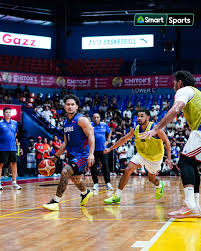 Basketball game in progress on an indoor court; players in blue and yellow jerseys chase the ball with spectators in the stands behind.