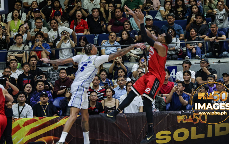 Two basketball players in white and red jerseys leap for the ball as a crowded arena watches from the stands behind them, capturing a fast-paced moment.