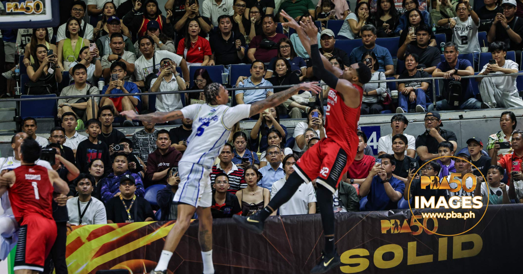 Two basketball players in white and red jerseys leap for the ball as a crowded arena watches from the stands behind them, capturing a fast-paced moment.