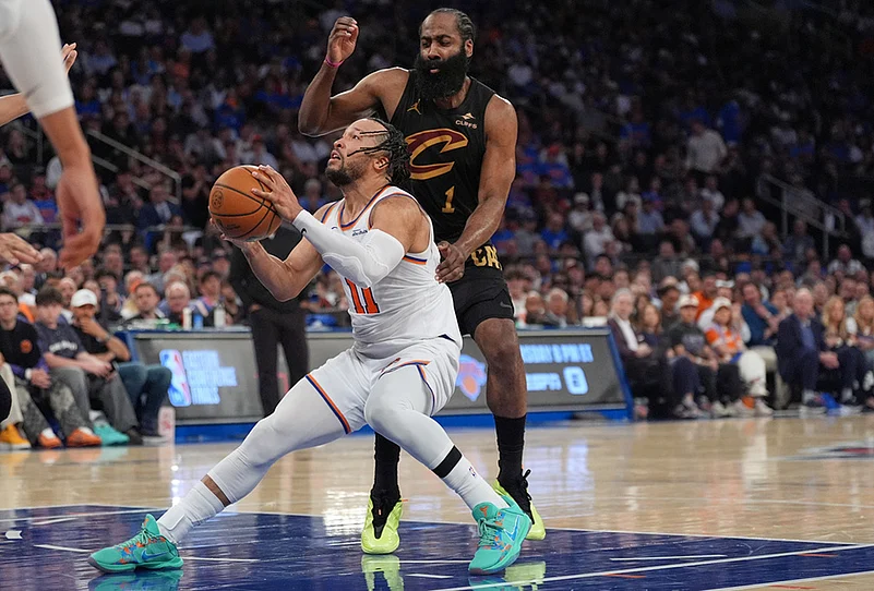 Knicks guard in white drives to the basket while a Cavaliers defender in black reaches from behind in a crowded arena.