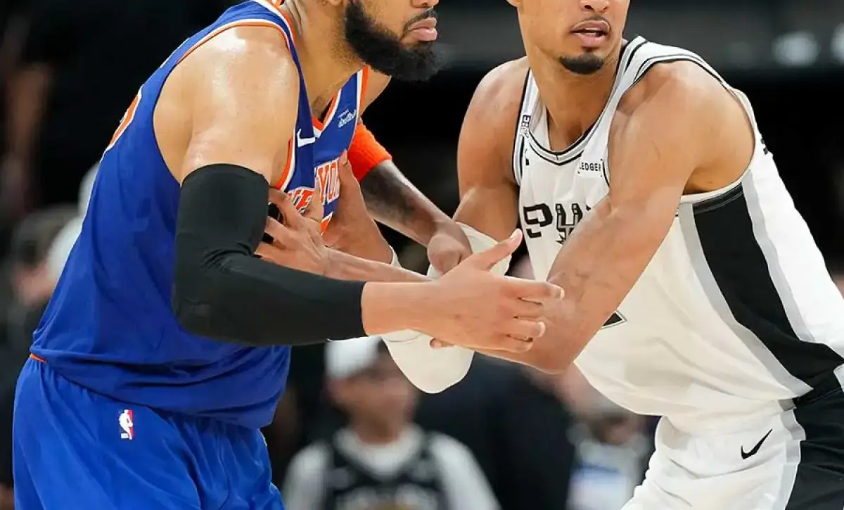 Two male basketball players in a hand-to-hand stance during a game: a Knicks player in blue and orange defending a Spurs player in white and black as spectators watch in the background.