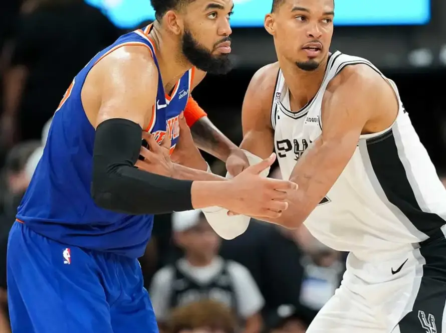 Two male basketball players in a hand-to-hand stance during a game: a Knicks player in blue and orange defending a Spurs player in white and black as spectators watch in the background.