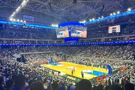 Full arena during a basketball game, packed with fans, bright court, and a scoreboard overhead.