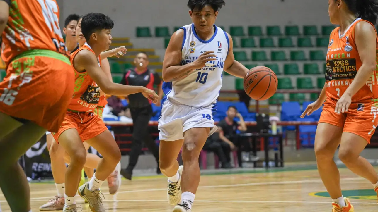 White-jersey player (number 10) dribbles toward the basket while orange-team defenders converge in a crowded, indoor basketball game.