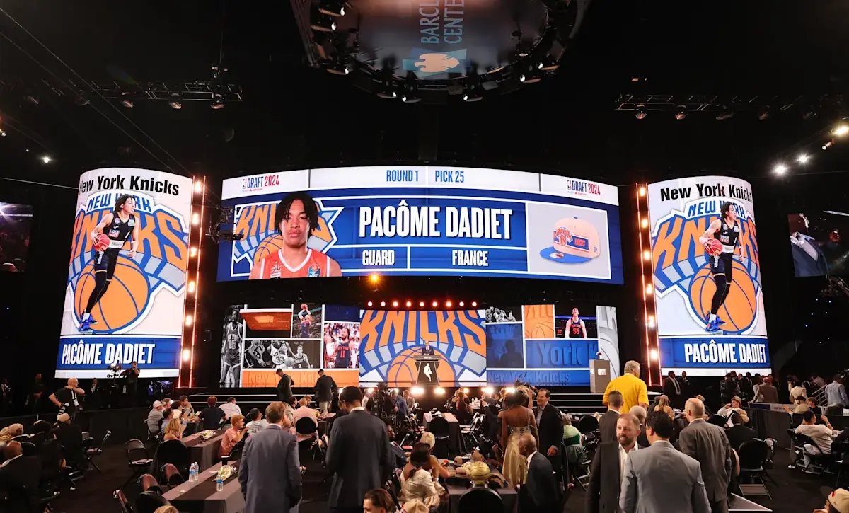 Large, illuminated basketball stage at the NBA Draft 2024 with Pacôme Dadiet's profile and 'New York Knicks' branding on surrounding screens, crowd seated and standing nearby.