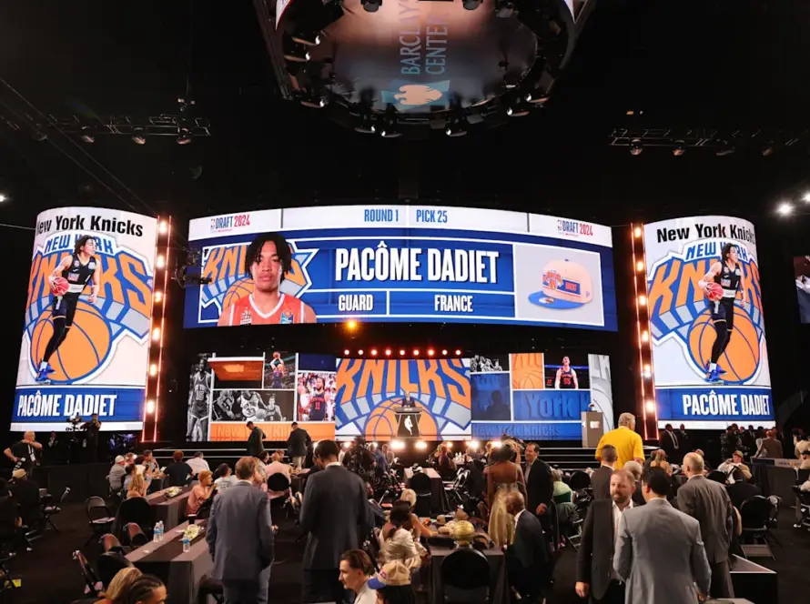 Large, illuminated basketball stage at the NBA Draft 2024 with Pacôme Dadiet's profile and 'New York Knicks' branding on surrounding screens, crowd seated and standing nearby.