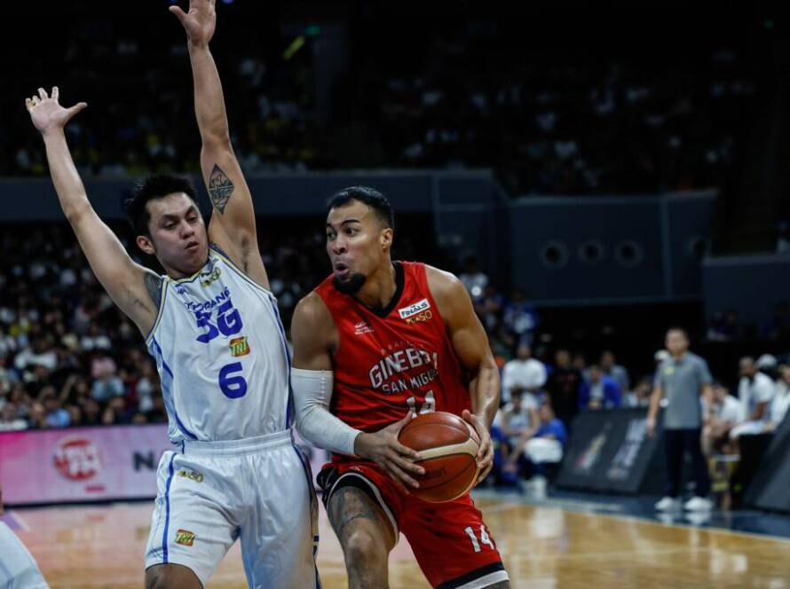 Basketball player in red dribbles toward the hoop while a defender in white jumps with arms raised to block the lane.