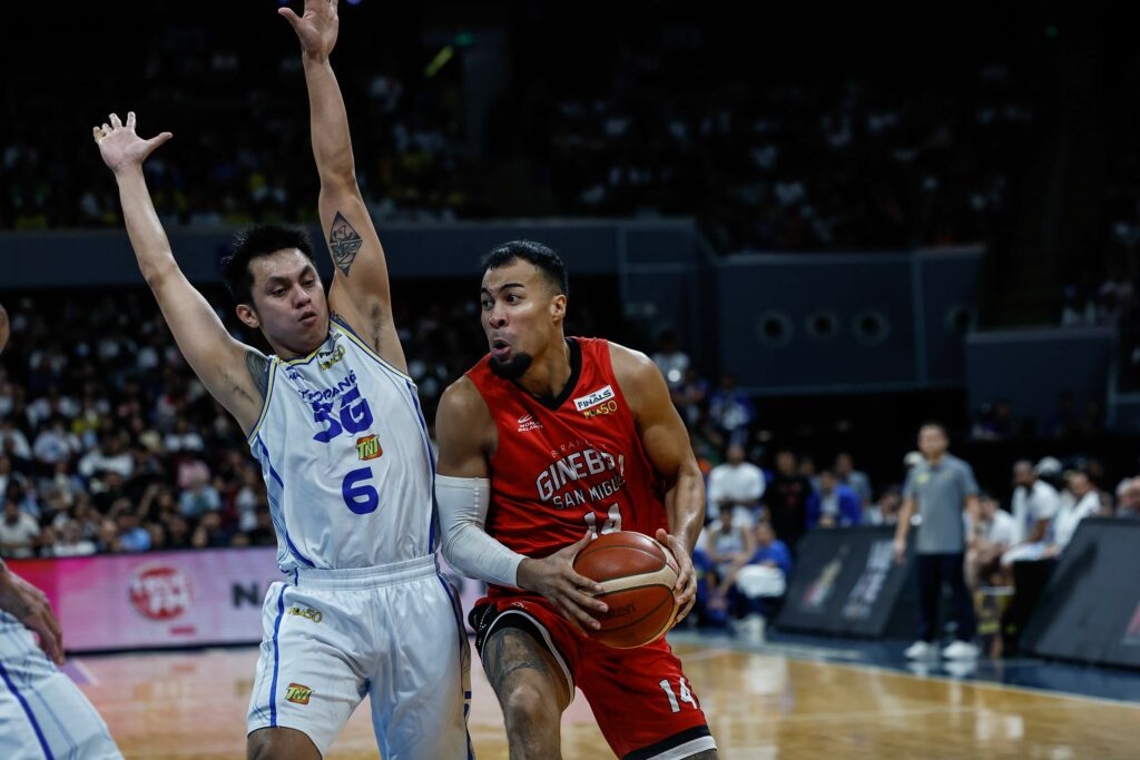 Basketball player in red dribbles toward the hoop while a defender in white jumps with arms raised to block the lane.