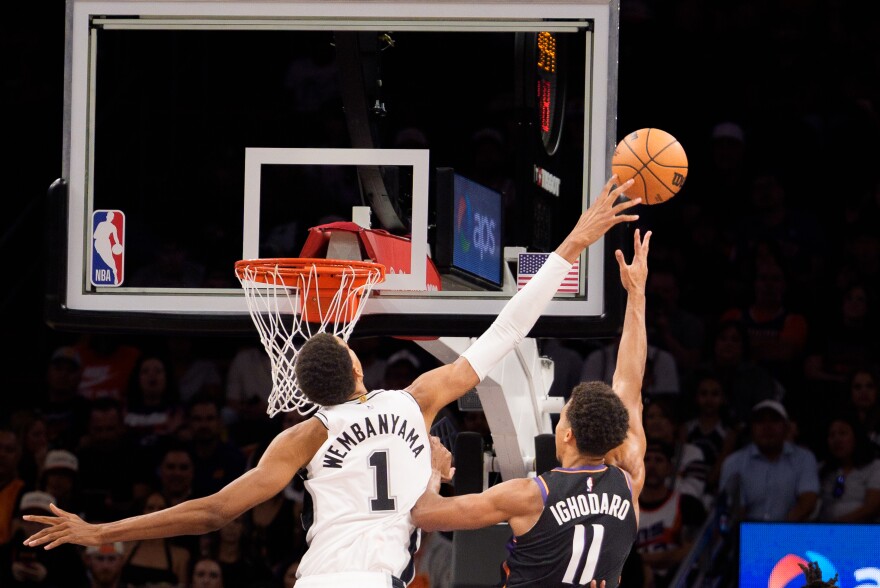 White-jersey player No. 1 reaches toward the basket while a defender No. 11 blocks the shot near the rim, with the backboard and net visible.