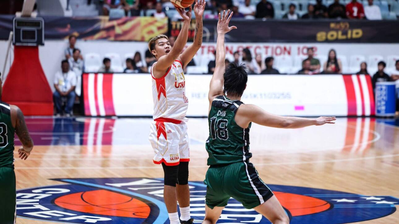 White-clad basketball player rises for a jump shot as a green-jersey defender reaches up to block in a crowded indoor arena.