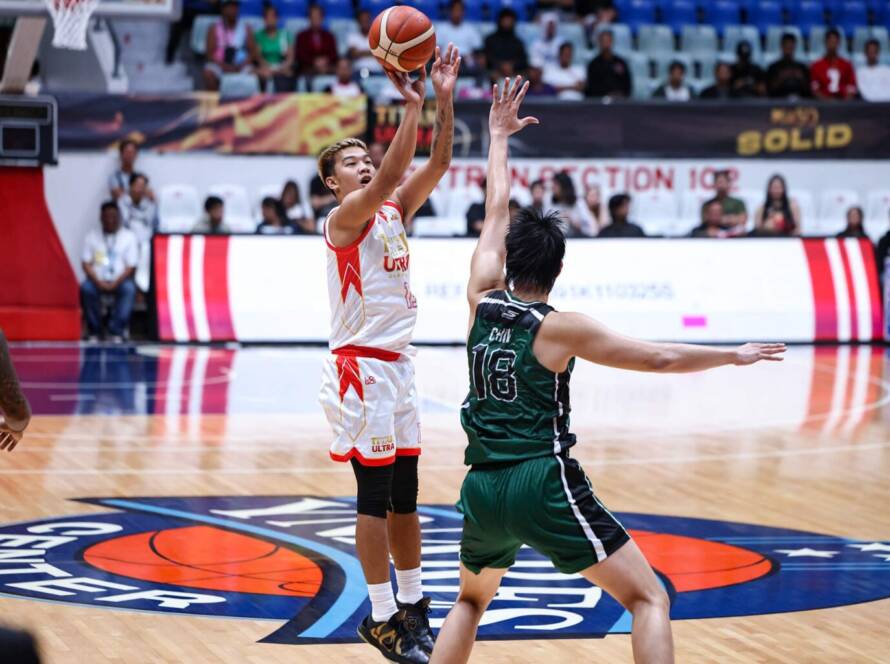 White-clad basketball player rises for a jump shot as a green-jersey defender reaches up to block in a crowded indoor arena.