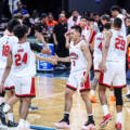 Basketball players in white and red jerseys high-fiving on a court after a game, with spectators in the stands behind them.