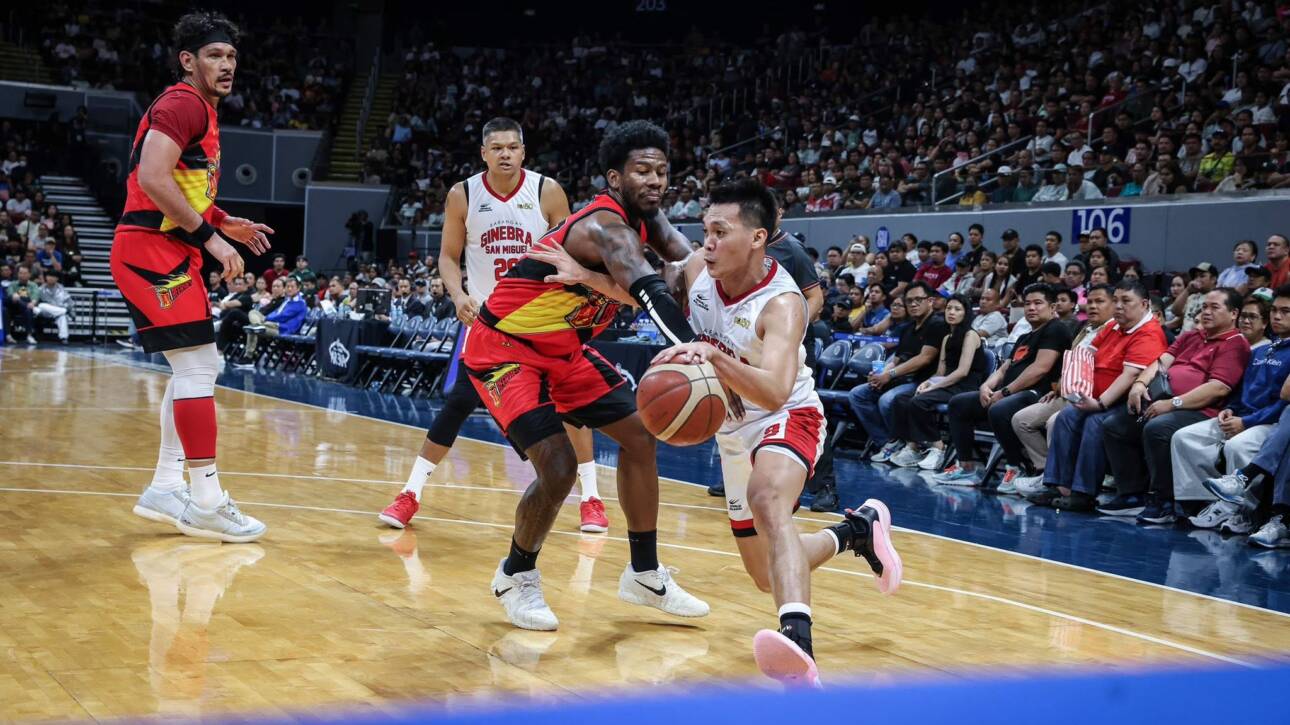 Basketball players clash near the baseline as one player drives with the ball while defenders converge and a packed arena watches.
