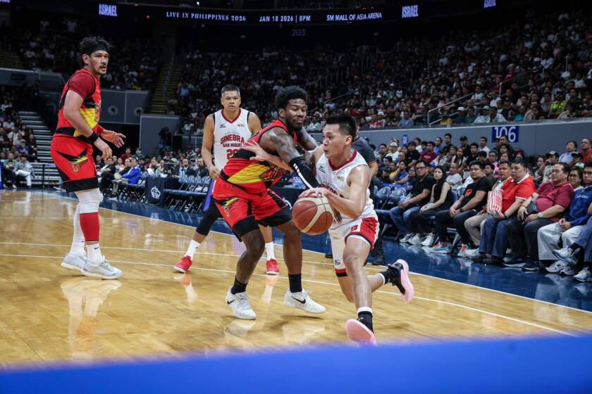 Basketball players clash near the baseline as one player drives with the ball while defenders converge and a packed arena watches.