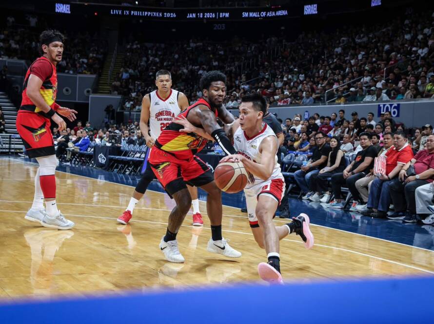 Basketball players clash near the baseline as one player drives with the ball while defenders converge and a packed arena watches.