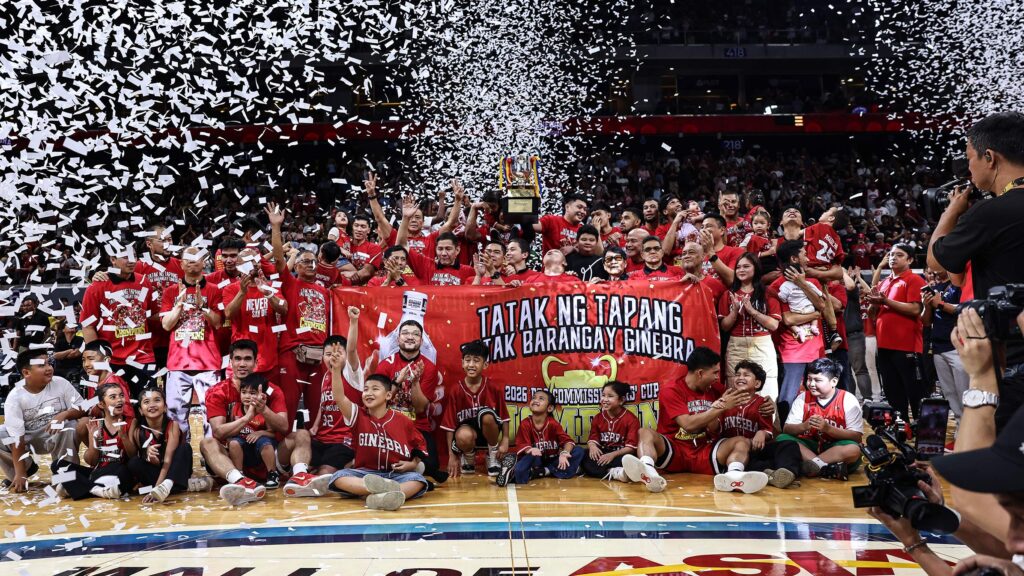 Basketball team and fans in red celebrate on court with a trophy and confetti filling the arena.