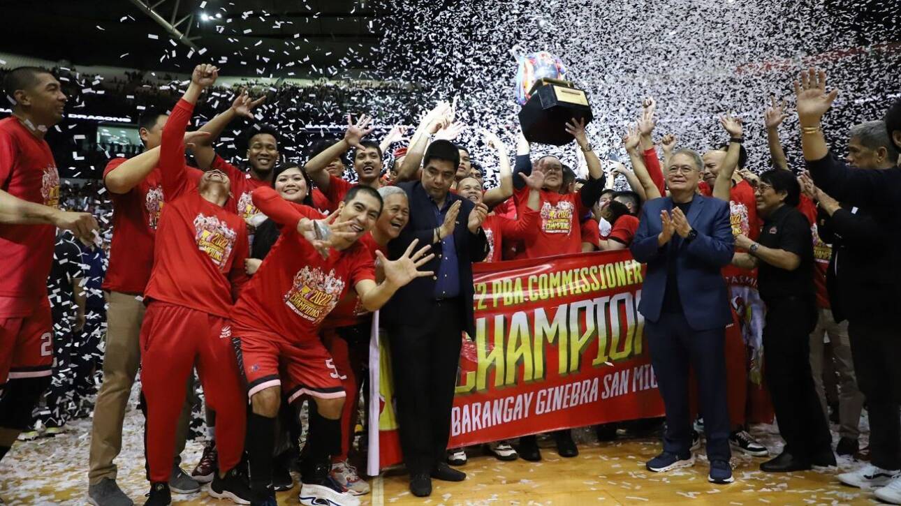Team in red uniforms celebrates winning a championship, raising a trophy amid confetti and cheering fans.