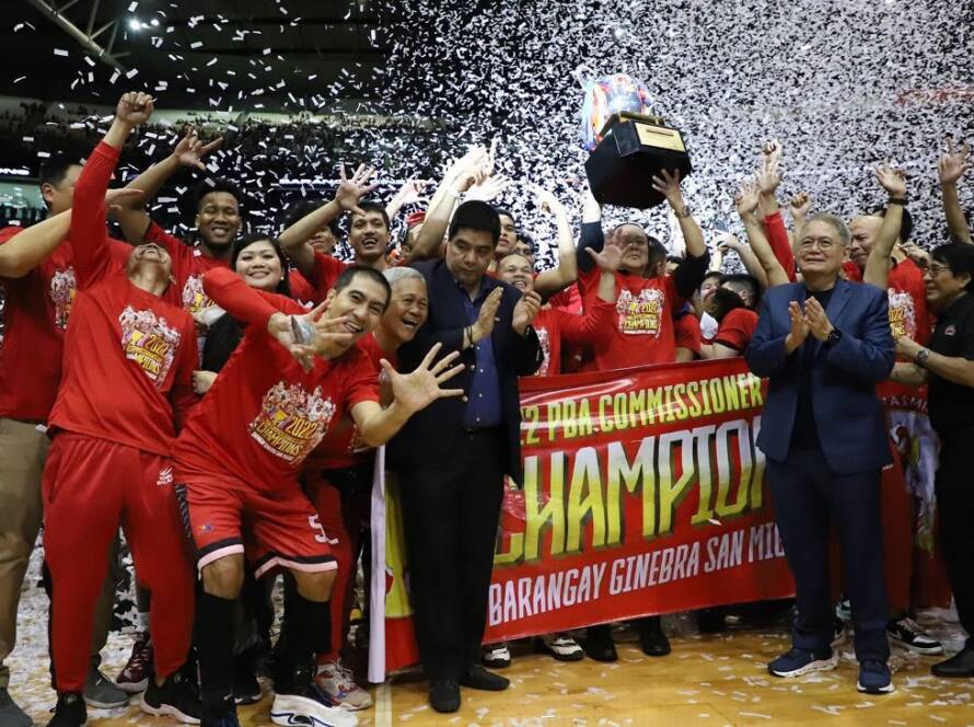 Team in red uniforms celebrates winning a championship, raising a trophy amid confetti and cheering fans.