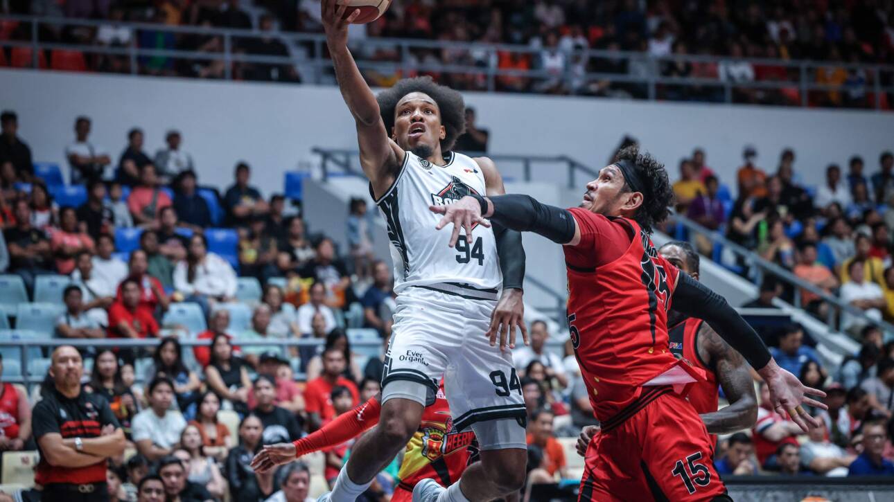 Basketball player in a white jersey leaps upward with the ball as a defender in red reaches in a crowded arena.