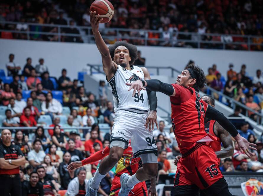 Basketball player in a white jersey leaps upward with the ball as a defender in red reaches in a crowded arena.