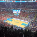 Wide-angle view of a crowded basketball arena with red and blue-clad fans watching a game and a large circular scoreboard above the court.