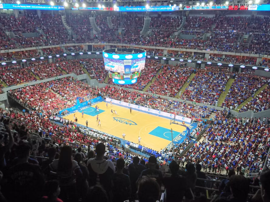 Wide-angle view of a crowded basketball arena with red and blue-clad fans watching a game and a large circular scoreboard above the court.