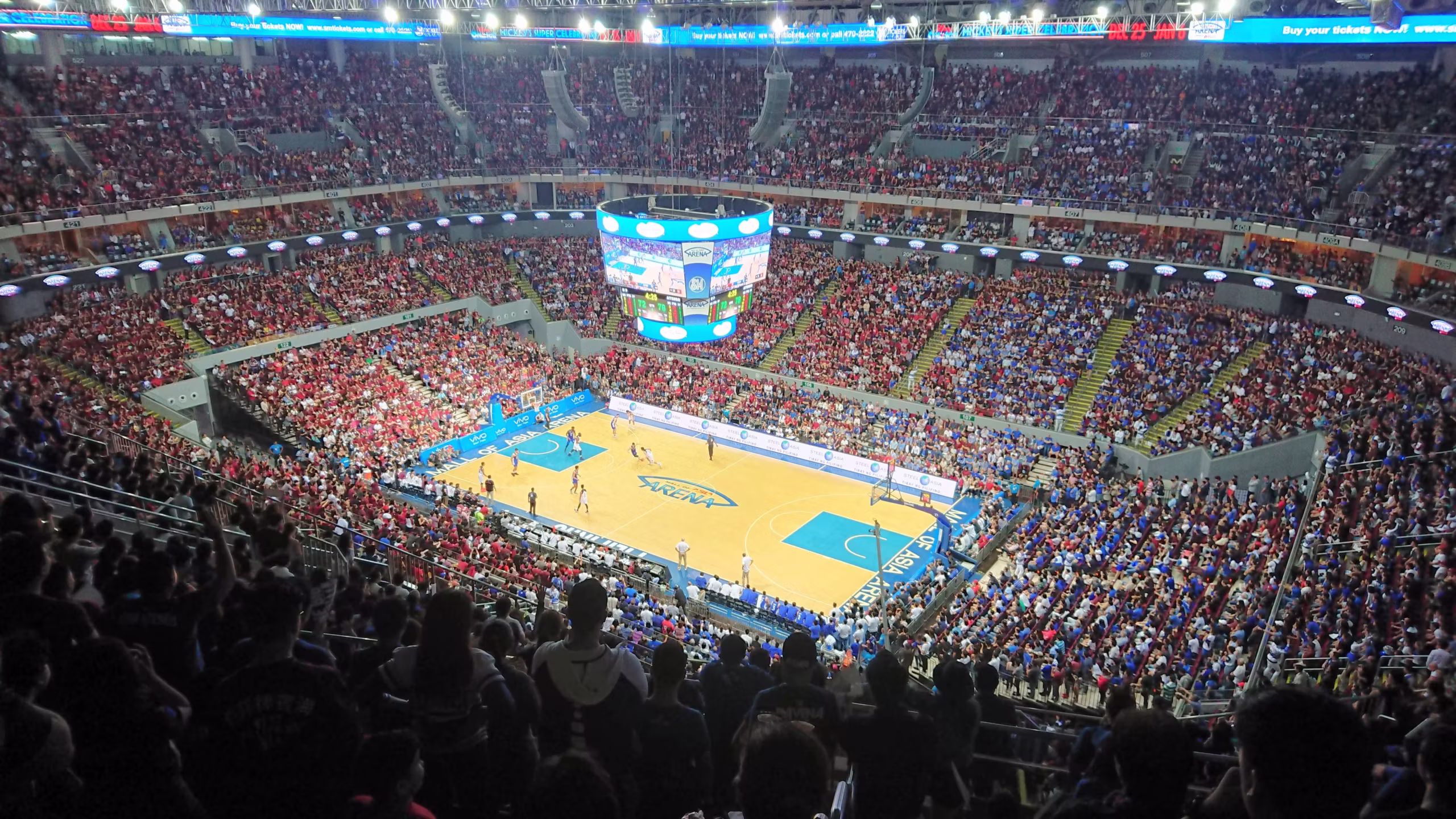 uaap-season-89-moa-arena-opening | GGSLOT | Sports & Games News Wide-angle view of a crowded basketball arena with red and blue-clad fans watching a game and a large circular scoreboard above the court.